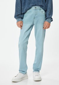 Vaqueros de mezclilla azul claro con corte recto, que cuentan con cinco bolsillos y costuras sutiles. Combinados con zapatillas blancas y una camiseta de manga larga azul.