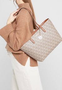 Sac cabas à motif beige avec des poignées en cuir marron, agrémenté d'un pendentif décoratif. Le sac a une forme structurée et une surface texturée.