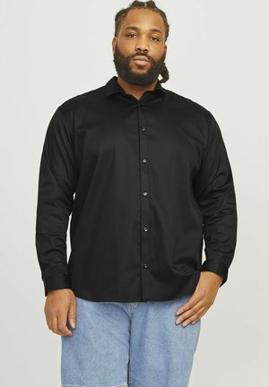 Camisa negra de manga larga con botón en el frente, hecha de una tela suave. Cuenta con un cuello clásico y un corte relajado, combinada con shorts de mezclilla claros.