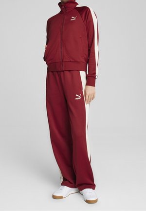 Osoba ubrana w bordowy dres Puma z białymi paskami po bokach i białe trampki, stojąca na tle jednolitego jasnoszarego tła.