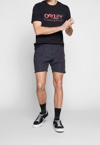 T-shirt noir avec logo "Oakley" rouge, shorts foncés et chaussures montantes noires ; le modèle présente une tenue athlétique et décontractée.
