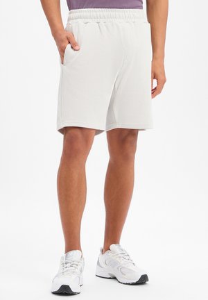 Mann trägt weiße gerippte Shorts mit elastischem Bund und Seitentaschen, kombiniert mit weißen Sportschuhen, steht vor weißem Hintergrund.