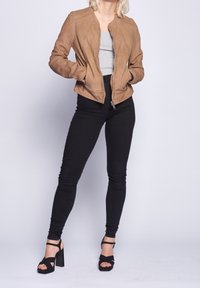 Braune Kunstsuede-Bomberjacke mit Reißverschluss vorne, hohem Kragen und diagonalem genähten Details, kombiniert mit schwarzen, engen Hosen und schwarzen Absätzen.