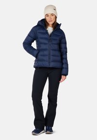 Marineblå pufferjakke med lynlås foran og hætte, kombineret med sorte bukser og grå sneakers. Modellen har en beige strikket hue på.