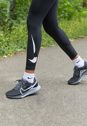 Person trägt schwarze Nike-Leggings, weiße Nike-Socken und schwarze Nike-Laufschuhe und läuft auf einem gepflasterten Weg mit Grünflächen in der Nähe.