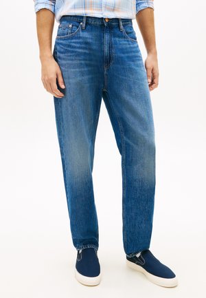 Mężczyzna w średnio-niebieskich prostych dżinsach, jasnoniebieskiej koszuli w kratę włożonej do środka oraz granatowych wsuwanych trampkach z białą podeszwą.