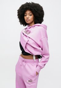 Ροζ φούτερ με κουκούλα και λεπτομέρεια λογότυπου, συνδυασμένο με ροζ φόρμα σετ. Το ύφασμα φαίνεται απαλό με ελαστικές μανσέτες και κορδόνια για ρυθμιζόμενη εφαρμογή.