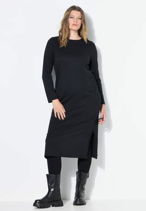 Zwarte lange mouw jurk met een zijslit, gemaakt van een gladde stof. Draagt met zwarte enkellaarsjes en leggings, en toont een eenvoudig, strak ontwerp.