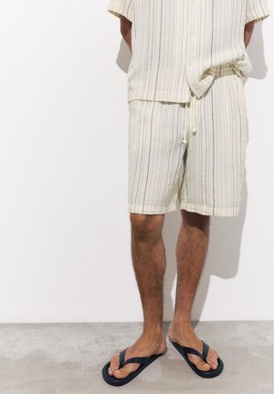 Homme portant un short beige rayé et une chemise assortie avec des tongs noires, debout contre un mur blanc sur un sol gris.