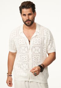 Weißes kurzärmliges Strickshirt mit aufwendigen spitzenähnlichen Mustern und einem Button-Down-Kragen. Auffällige Textur und detaillierte Verarbeitung.