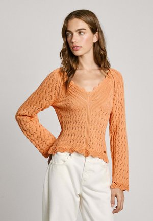En orange strikket cardigan med en scalloped halsudskæring og bølget tekstur. Lange ærmer og en tætsiddende silhuet, parret med hvide bukser.