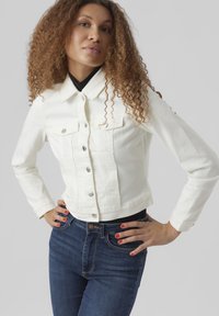 Veste en denim blanc avec un col, fermeture à boutons sur le devant et deux poches poitrine. Associée à un jean bleu foncé. Texture lisse.