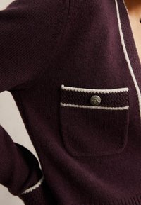 Cardigan bordeaux cu un tricotaj texturat, având un buzunar pe piept cu bordură albă și un nasture decorativ, precum și manșete cu model rib. 