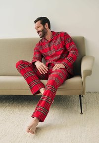 Rød ternet pyjamas sæt med en knap-op bluse med krave og matchende bukser. Blødt stof med en hyggelig pasform, båret mens man sidder på en sofa.