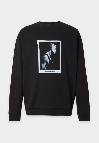 Sweat-shirt noir à manches longues avec une photo de profil en noir et blanc centrée d’un homme dans un cadre blanc et un texte "EMPORIO ARMANI" en dessous.