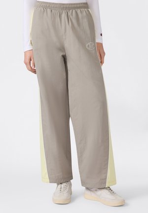 Pantaloni bej cu picioare largi, cu panouri laterale galben pal, talie elastică și logo conturat alb pe coapsa stângă, purtați cu adidași albi.