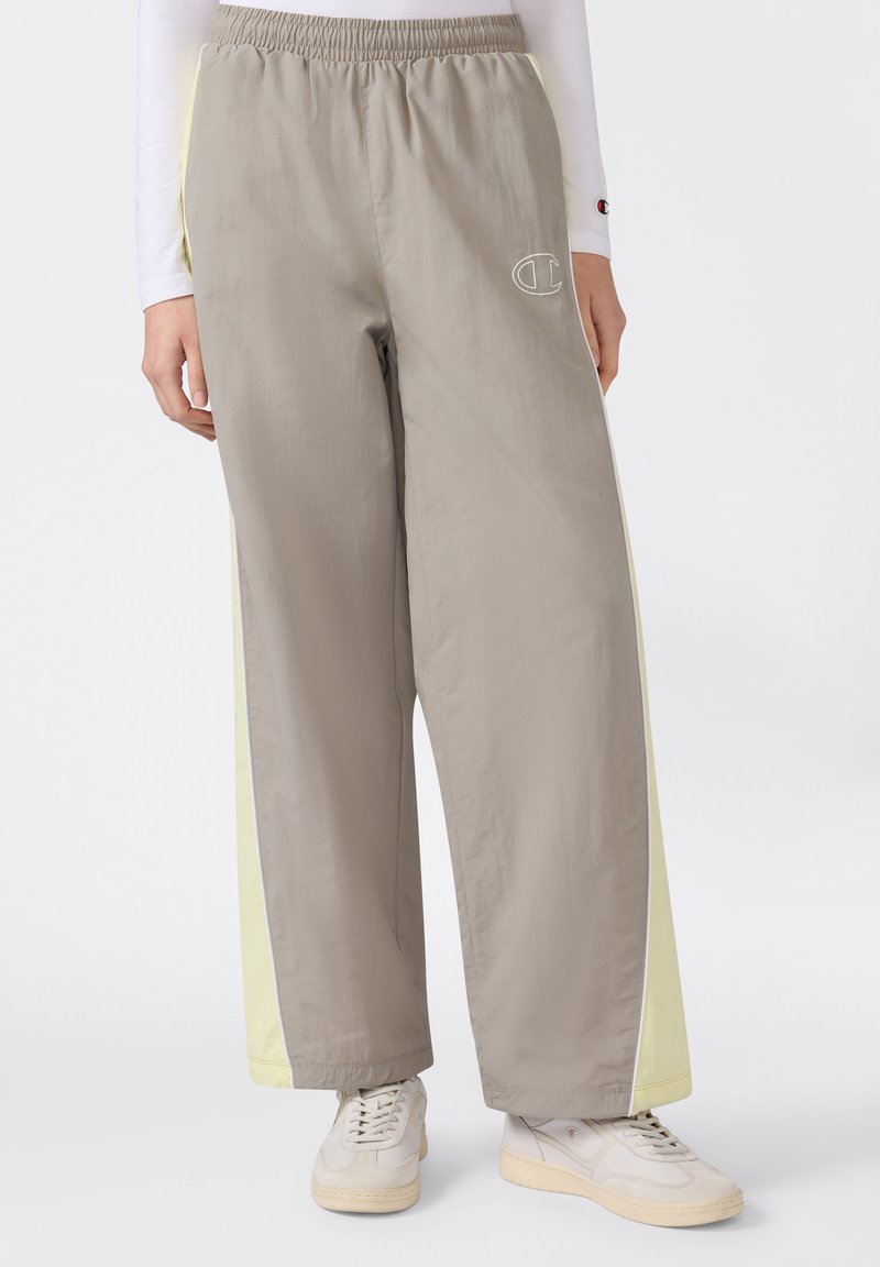 Μπεζ παντελόνι με φαρδιά μπατζάκια και ανοιχτοκίτρινες πλαϊνές λωρίδες, ελαστική μέση και λευκό περίγραμμα λογότυπου στο αριστερό μηρό, φορεμένο με λευκά αθλητικά παπούτσια.