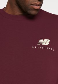 Primer plano de una persona que lleva una camiseta de cuello redondo color burdeos con un logo blanco de "NB Basketball" en el pecho.