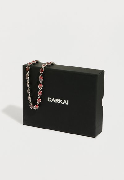 Srebrna verižica z ovalnimi rdečimi dragimi kamni, prekrita čez črno škatlo z znamko DARKAI na belem ozadju.