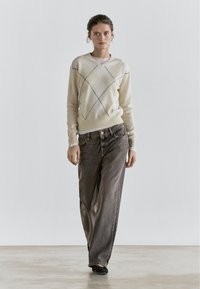 Cream sweater with diamond pattern and black accents, paired with loose dark gray jeans. Model stands on a neutral background.