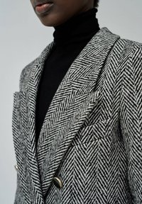 Blazer a fantasia houndstooth in bianco e nero con finitura testurizzata, dotato di un design doppiopetto e dettagli in metallo dorato.