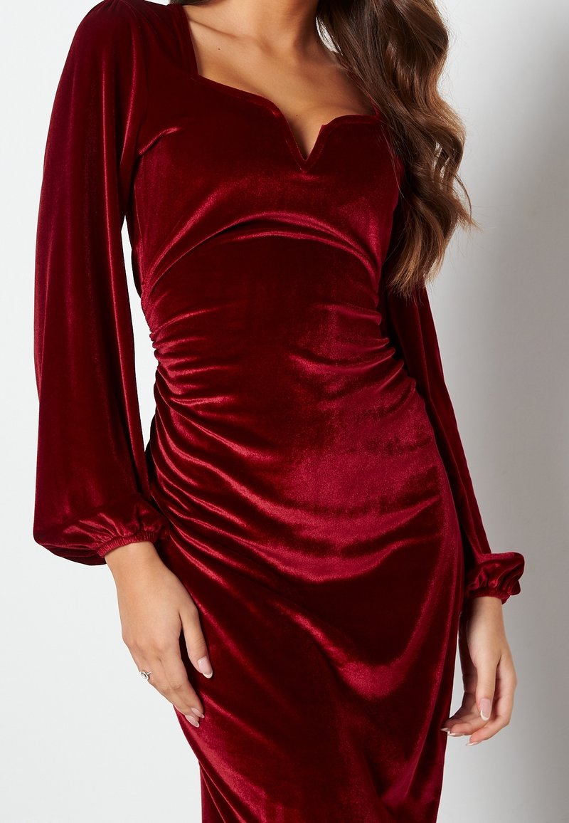Femme portant une robe en velours rouge foncé cintrée à manches longues, avec un décolleté en V et des fronces à la taille.