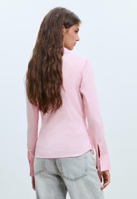 Camicia a maniche lunghe rosa chiaro con righe verticali, dotata di colletto e polsini con spacco, abbinata a jeans grigi con texture.