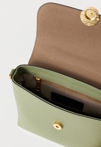 Πράσινη τσάντα από υφή δέρματος με κλεισίματος με χρυσό κουμπί, διαθέτει μπεζ εσωτερικό και τσέπη με φερμουάρ. Ανοικτή για να εμφανιστούν τα εσωτερικά διαμερίσματα.