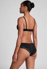 Mujer con el cabello recogido en un moño, vistiendo un sujetador negro y una braguita negra con encaje, de perfil contra un fondo sencillo.