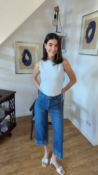 Kvinne smiler i hvit ermeløs topp, korte blå jeans med bølgete kanter og hvite åpne hæler i et moderne rom med innrammet kunst.