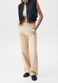 Beige weit geschnittene Hose mit Taschen auf der Vorderseite, kombiniert mit einer schwarzen ärmellosen Jacke in Cropped-Länge und schwarzen Sandalen. Glatter Stoff mit lässigem Schnitt.