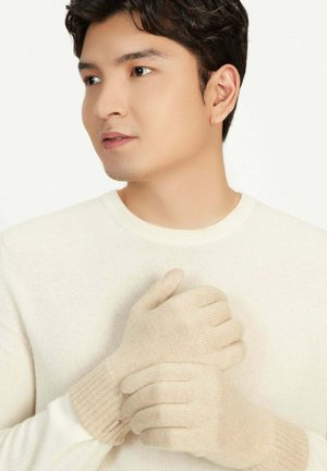 Junger Mann mit cremefarbenen Handschuhen und passendem Pullover, der zur Seite schaut, vor einem einfarbigen hellen Hintergrund.