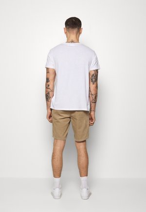Camiseta blanca de cuello redondo, pantalones cortos de pana color caqui y zapatillas blancas. Tatuajes visibles en los brazos. Fondo sencillo. Atuendo casual.
