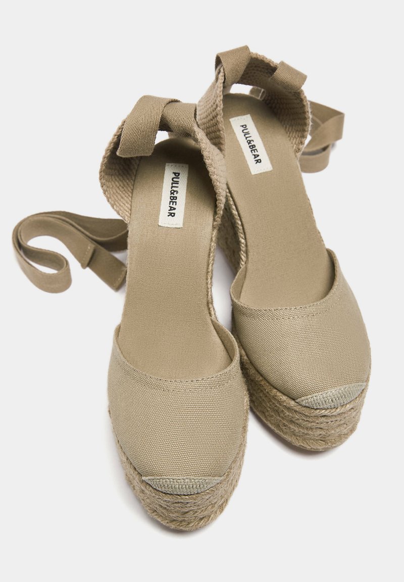 Beige Canvas-espadrilles mit einer gewebten Jutesohle, runder Zehenpartie und einem Umschlagriemen um den Knöchel. Flache Textur mit einem kontrastierenden Zehenkappen-Detail.