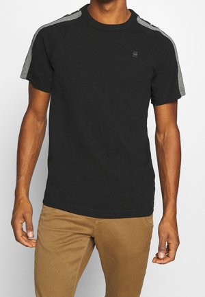 Homme portant un T-shirt noir à manches courtes avec des rayures grises sur les manches et un petit logo sur la poitrine, associé à un pantalon marron clair.