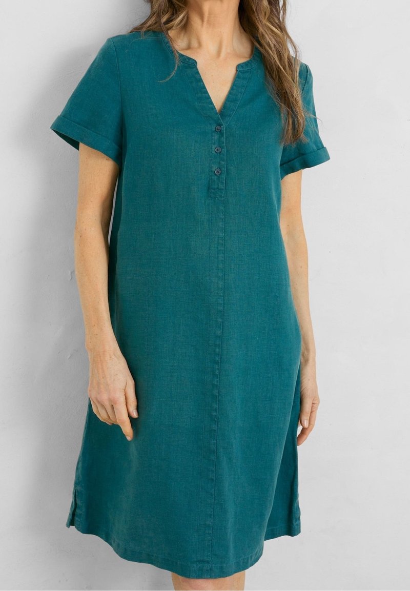 Vestido de lino en tono verde azulado con mangas cortas con vuelta, escote en V y botonera. Corte recto hasta la rodilla con aberturas laterales para mayor movilidad.