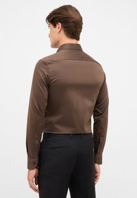 Braunes langärmeliges Hemd mit glatter Textur, klassischem Kragen und geknöpften Manschetten; kombiniert mit schwarzen Hosen mit Gesäßtaschen.