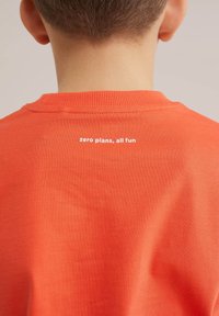 Enfant portant un t-shirt orange avec le texte « zero plans, all fun » imprimé dans le haut du dos, tête tournée loin de la caméra.