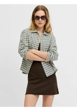 Kobieta z długimi blond włosami, nosząca ciemne okulary przeciwsłoneczne, koszulę w kratę z podwiniętymi rękawami i krótką ciemną sukienkę, z założonymi rękami.