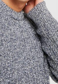 Strickpullover in Blau- und Grautönen, mit geripptem Muster und hohem Kragen. Nahaufnahme des Ärmeldetails.