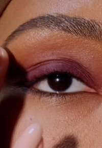 Gros plan sur un œil avec un iris marron foncé et une paupière arborant un fard à paupières bourgogne profond. Texture de peau lisse et détails des sourcils bien marqués.
