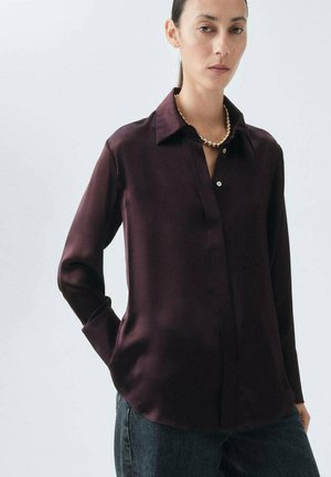 Blusa di seta borgogna con colletto, bottoni frontali, maniche lunghe e orlo leggermente curvato, abbinata a jeans scuri.