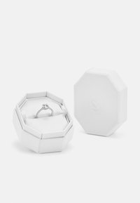 Achteckige weiße Schmuckschatulle mit strukturierter Oberfläche, die einen silbernen Ring mit einem zentralen Stein und kleineren Akzentsteinen, ebenfalls aus Silber, enthält.