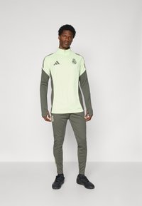 Helles grünes Sportoberteil mit Reißverschluss, grauen Akzenten und Teamlogo; kombiniert mit dunkelgrauen Jogginghosen mit seitlichen Streifen und körpernahem Design.