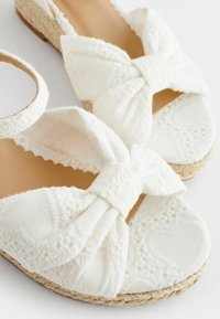 Sandali espadrillas bianchi con un fiocco in tessuto, bordi ondulati, motivi floreali ricamati e una suola avvolta in juta.