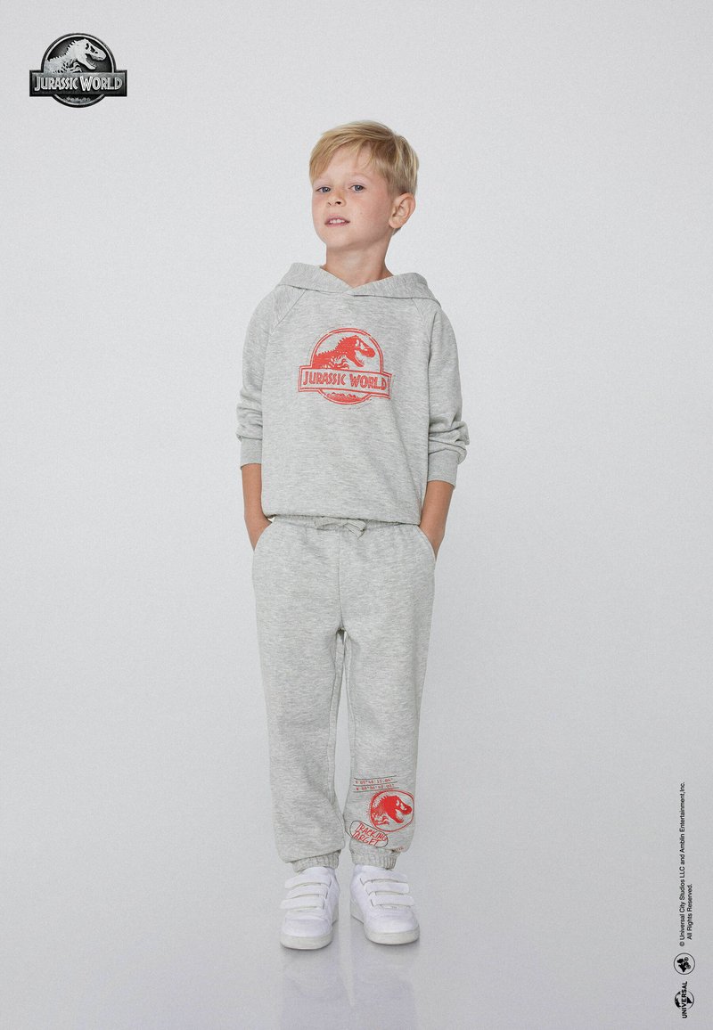Szara bluza z kapturem z czerwonym logo Jurassic World, w połączeniu z dopasowanymi spodniami dresowymi z akcentem logo. Materiał wydaje się miękki i casualowy.