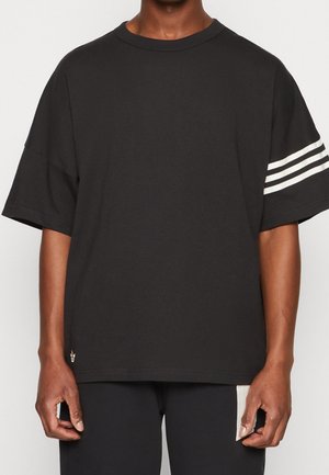 Person trägt ein schwarzes T-Shirt mit kurzen Ärmeln, drei weißen Streifen am linken Ärmel und einem kleinen Logo nahe dem unteren Saum.