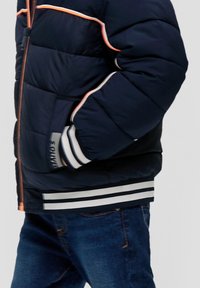 Marineblauwe gewatteerde jas met oranje en witte gestreepte accenten, ribbelmanchetten en een rits. Bevat een logo-label op de zijzak.