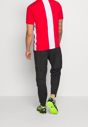 Mann, der weggeht und ein rot-weiß gestreiftes Hemd mit vertikalen Streifen, schwarze Hose, schwarze Sneaker mit neongrüner Sohle und sichtbare Arm-Tätowierungen trägt.