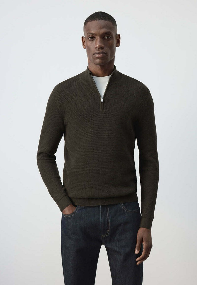 Homme portant un pull vert foncé à fermeture éclair quart, par-dessus une chemise blanche et un jean bleu foncé, debout devant un fond clair uni.